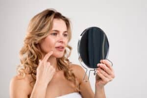 Mature woman looking at skin in mirror thinking about getting facetite or facelift.